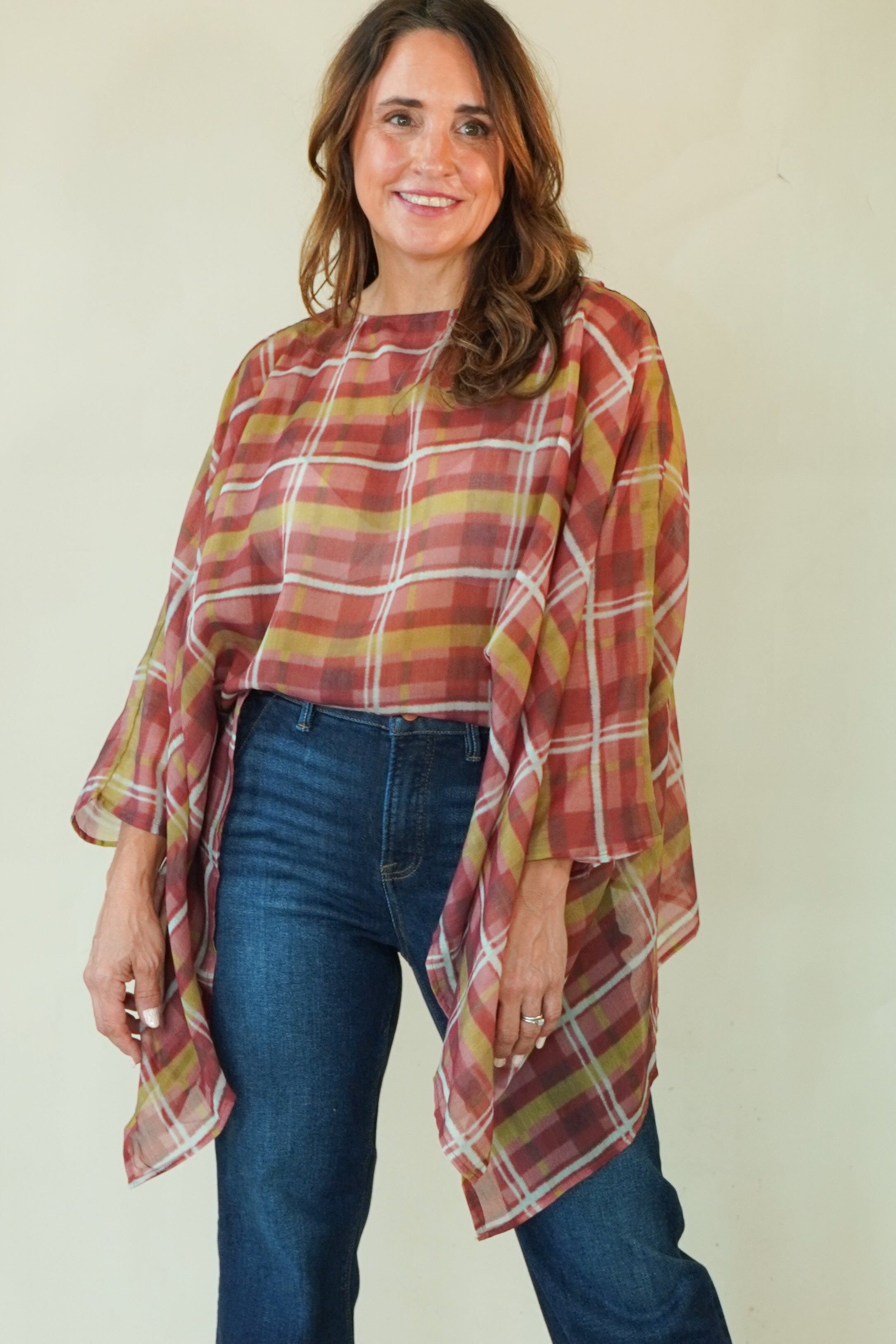 A woman modeling a rust-orange and yellow plaid poncho-style top with blue jeans and brown boots, standing against a plain light beige studio background.
