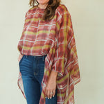 A woman modeling a rust red and mustard yellow plaid poncho-style top with blue jeans and brown boots, standing against a plain cream studio background.