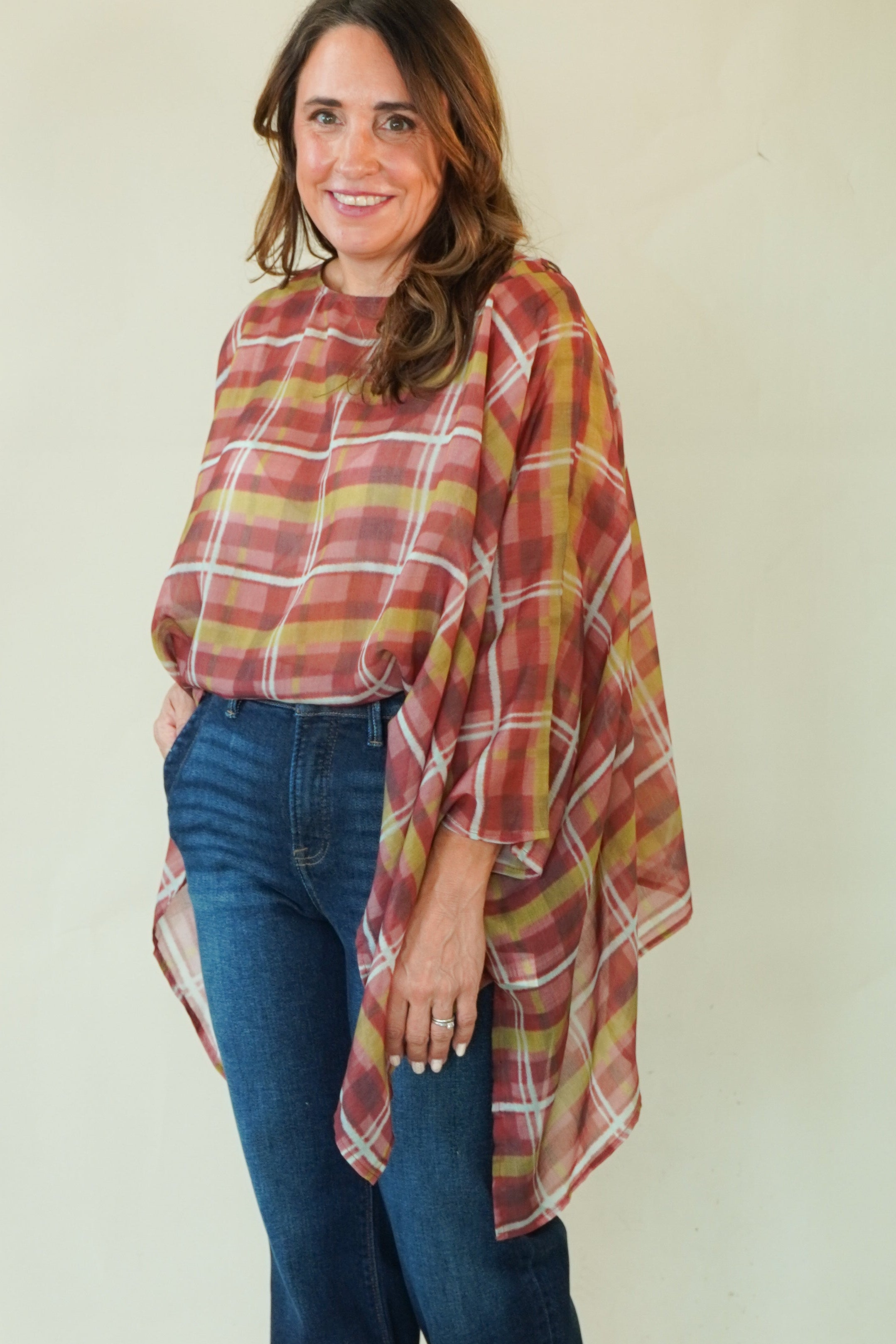 A woman modeling a rust red and mustard yellow plaid poncho-style top with blue jeans and brown boots, standing against a plain cream studio background.