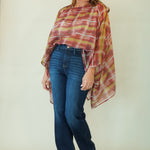 A woman modeling a rust-orange and yellow plaid poncho-style top with blue jeans and brown shoes, standing against a plain light beige studio background.