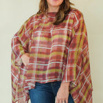 A woman modeling a rust red and mustard yellow plaid poncho top with blue jeans and brown boots, standing against a plain light beige background.