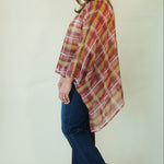 A woman modeling a rust red and mustard yellow plaid poncho-style top with blue jeans and brown leather cowboy boots, standing against a plain off-white studio background.