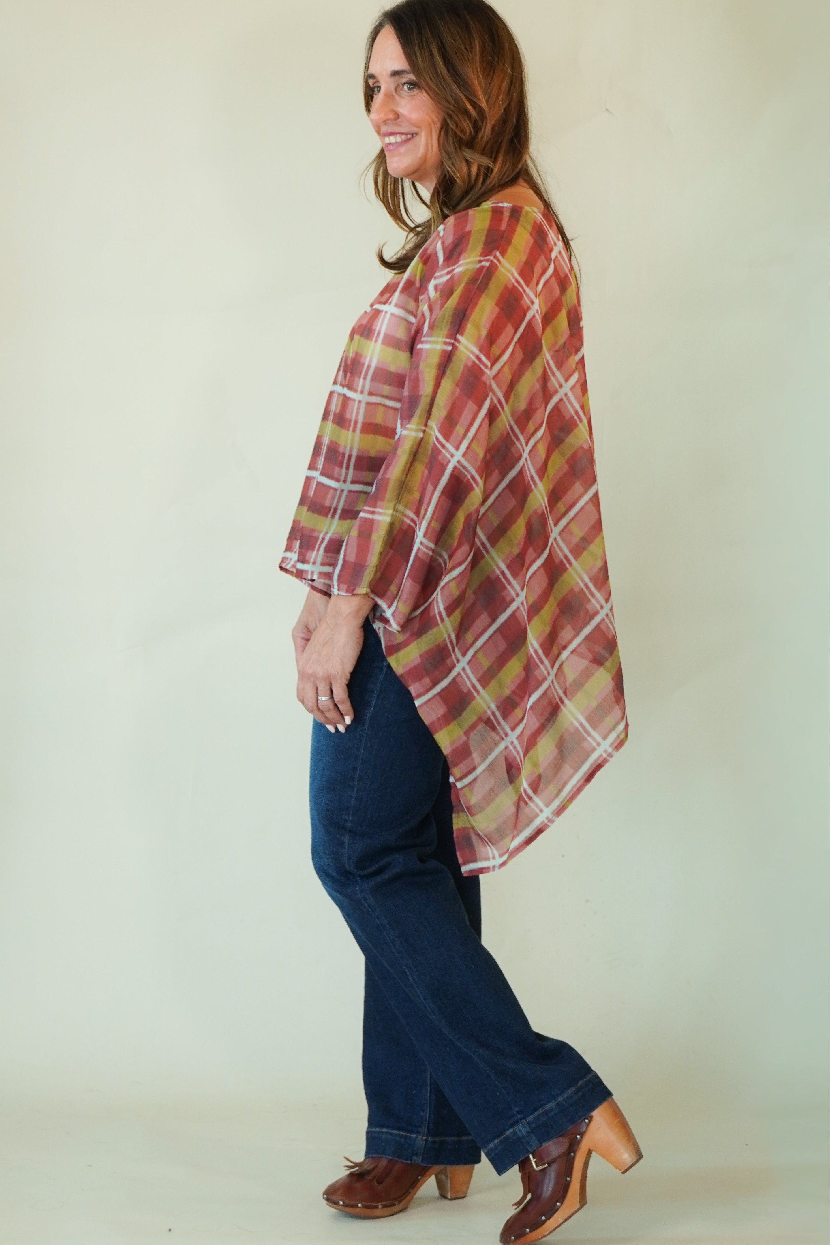 A woman modeling a rust red and mustard yellow plaid poncho-style top with blue jeans and brown leather cowboy boots, standing against a plain off-white studio background.