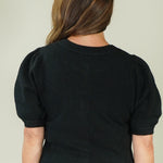 A woman viewed from the back wearing a black short-sleeve knit sweater with puffed shoulders and a ribbed crew neck, hem, and cuffs, paired with white pants against a plain light beige background.
