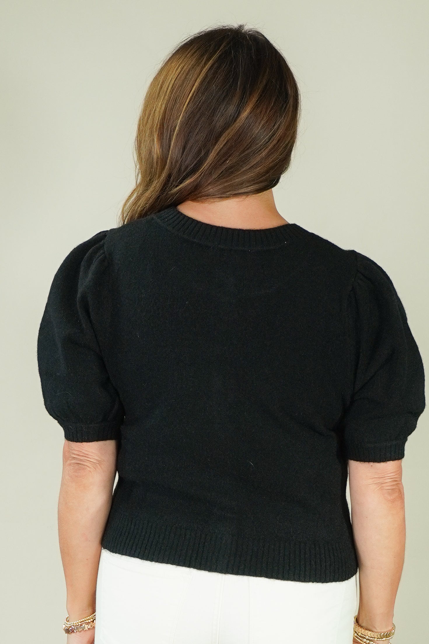 A woman viewed from the back wearing a black short-sleeve knit sweater with puffed shoulders and a ribbed crew neck, hem, and cuffs, paired with white pants against a plain light beige background.
