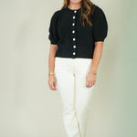A woman modeling a black short-sleeve cropped top with white buttons and white pants, standing against a plain light beige studio background.
