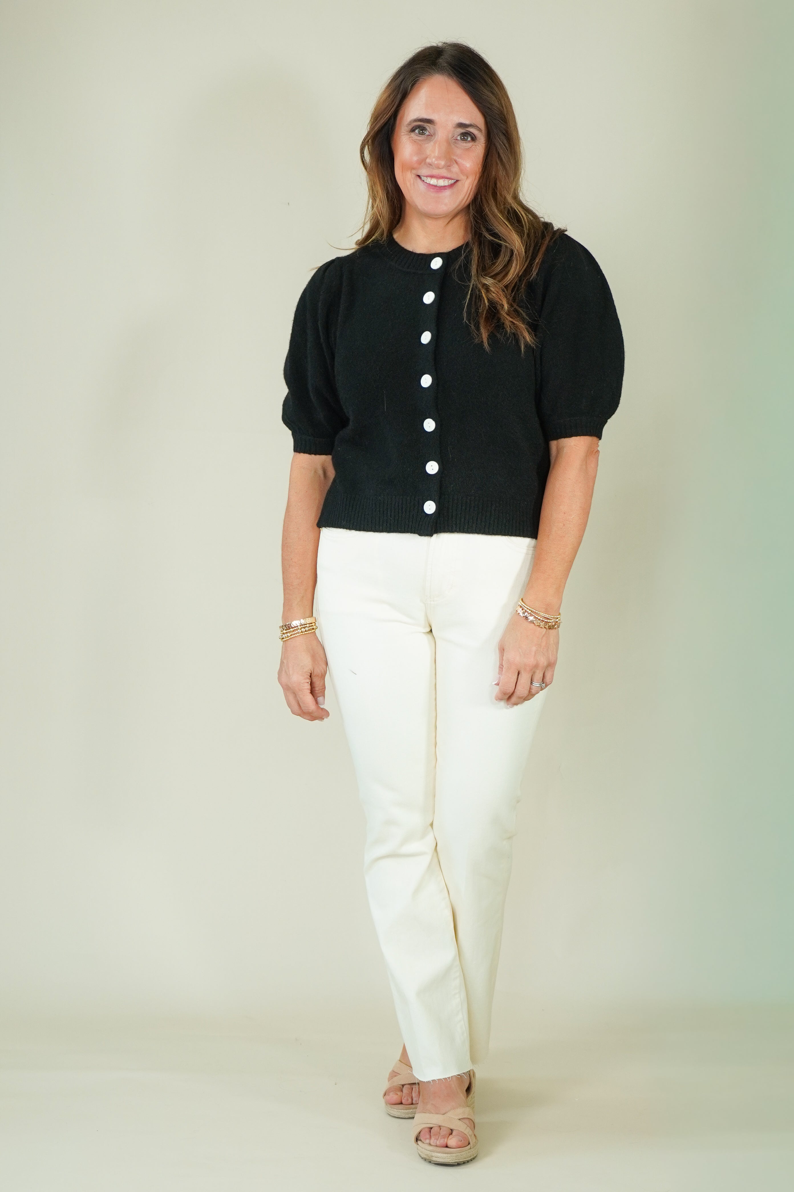 A woman modeling a black short-sleeve cropped top with white buttons and white pants, standing against a plain light beige studio background.