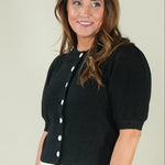 A woman modeling a short-sleeve black knit top with white buttons and white pants, standing against a plain light beige studio background.