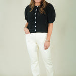 A woman modeling a black short-sleeve knit top with white buttons and white pants, standing against a plain light beige studio background.