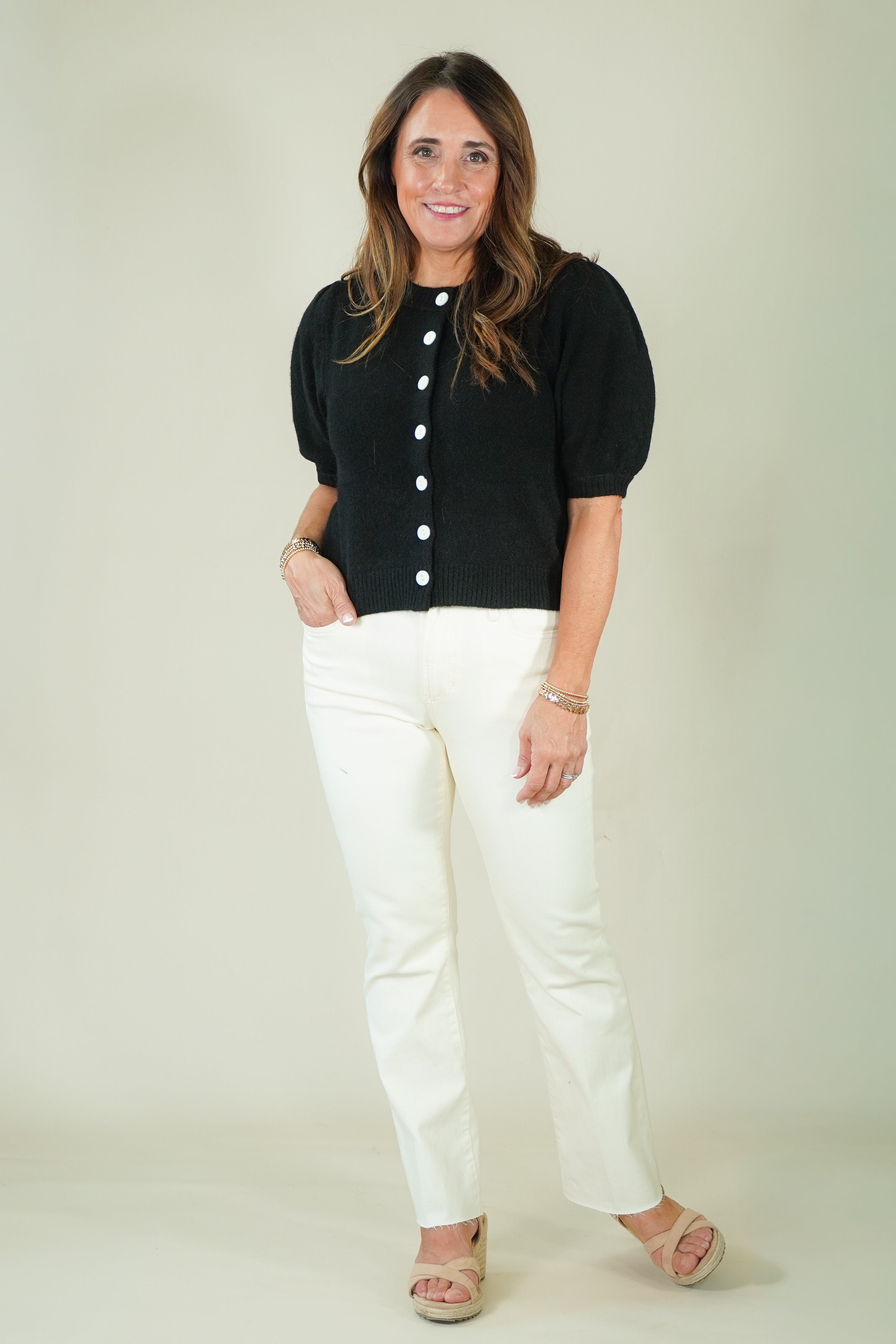 A woman modeling a black short-sleeve knit top with white buttons and white pants, standing against a plain light beige studio background.