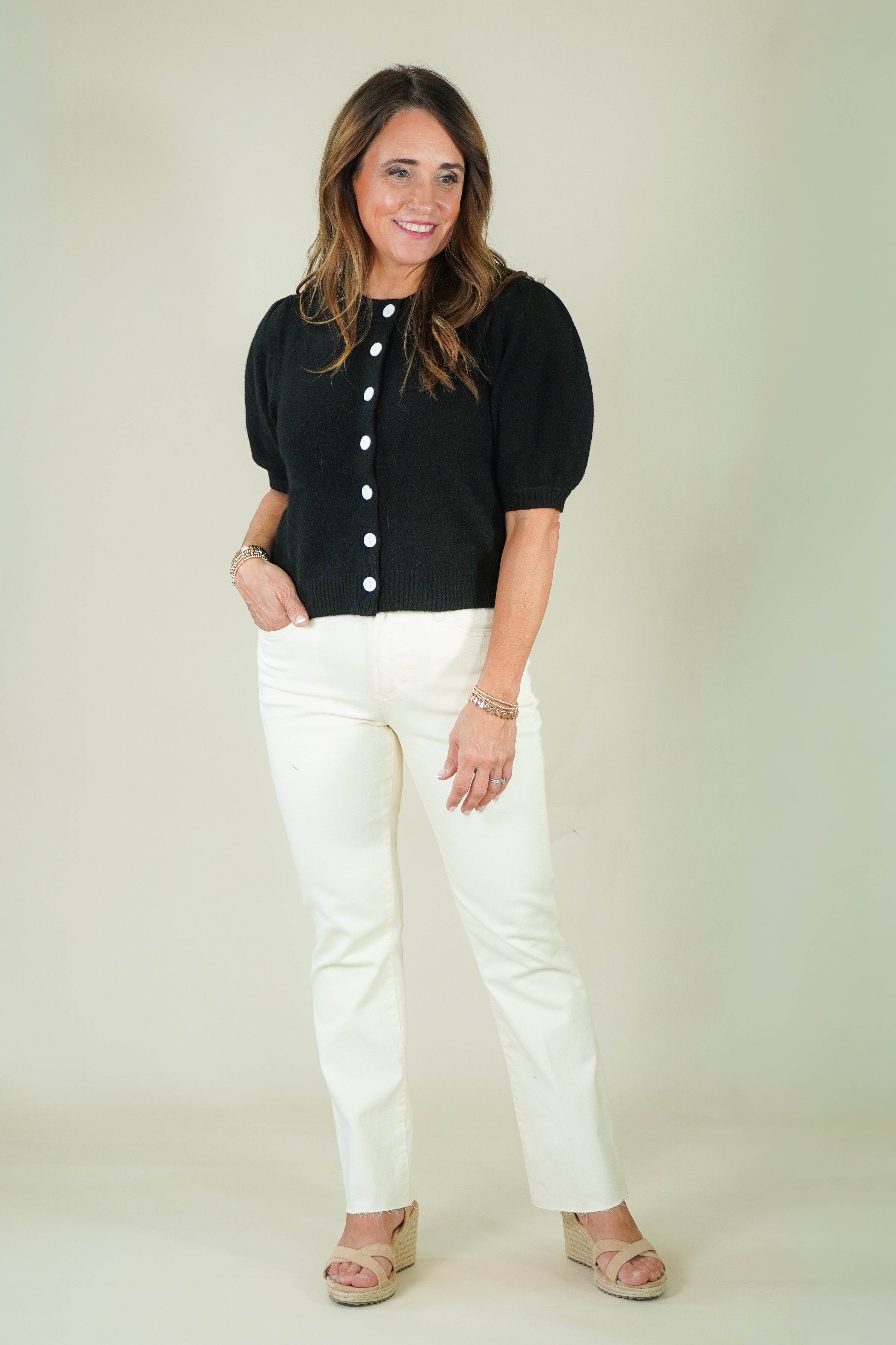 A woman modeling a black short-sleeve puff-sleeve top with white buttons down the front, paired with off-white straight-leg jeans and beige wedge sandals, against a plain light beige studio background.