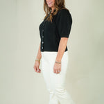 A woman modeling a black short-sleeve button-front cardigan with white pants and beige wedge sandals, standing against a plain light beige studio background.