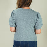 A woman viewed from behind wearing a heathered light blue short-sleeve knit top with puffed sleeves and dark blue jeans, against a plain light beige studio background.