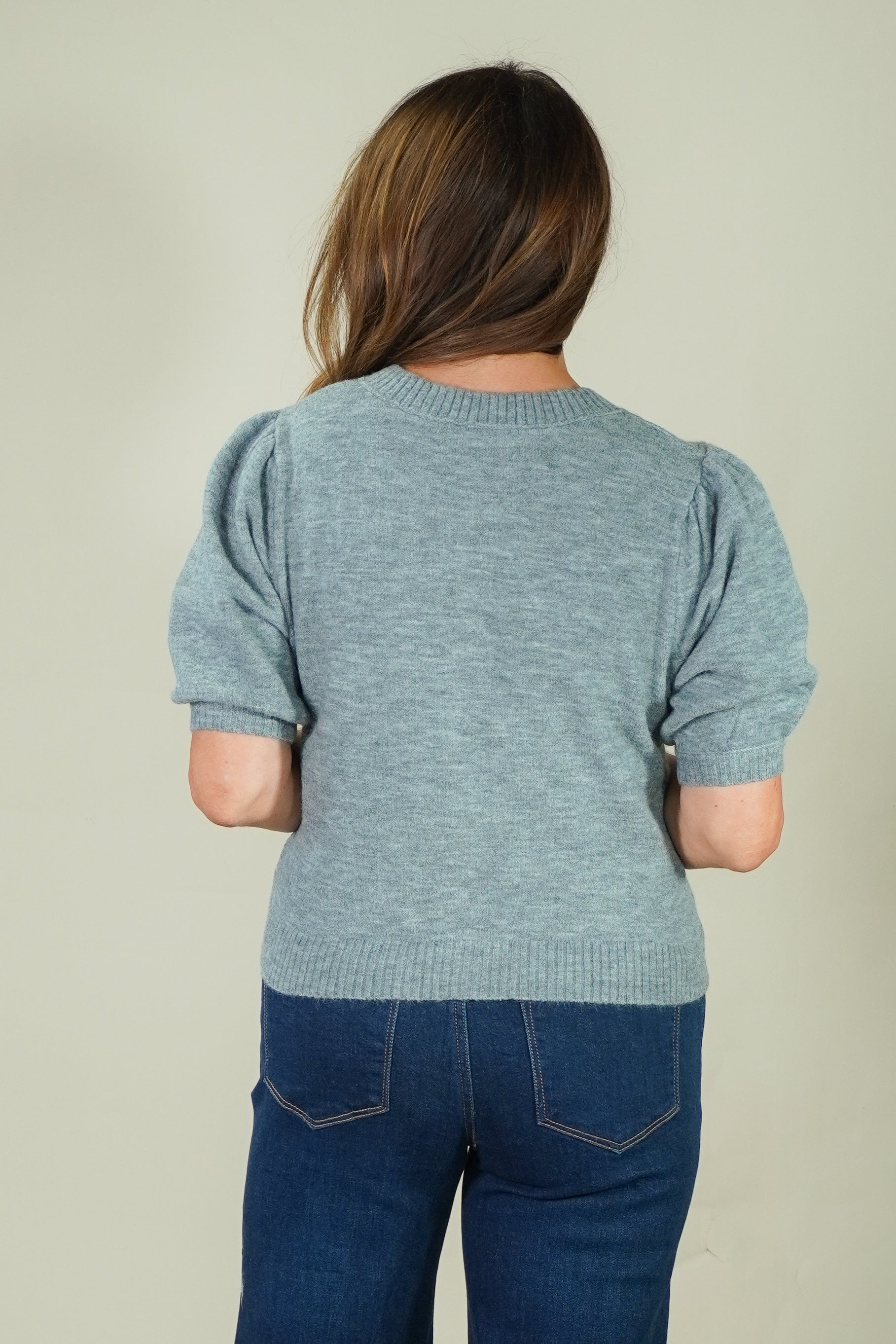 A woman viewed from behind wearing a heathered light blue short-sleeve knit top with puffed sleeves and dark blue jeans, against a plain light beige studio background.