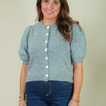 A woman wearing a light heather gray short-sleeve knit cardigan with white buttons and dark blue jeans, standing against a solid light beige background.