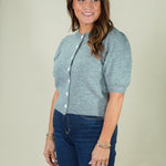A woman wearing a light heather gray short-sleeve knit cardigan with white buttons and dark blue jeans, standing against a solid light beige background.