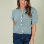 A woman modeling a light gray short-sleeve knit cardigan with puff sleeves and white buttons, paired with dark blue jeans, against a solid light beige background.