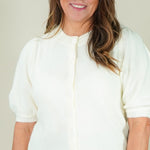 A woman modeling a cream-colored short-sleeve knit top with puff sleeves and a button-up front, paired with dark blue jeans, against a plain beige studio background.