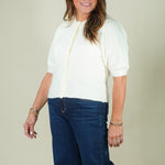 A woman modeling a cream short-sleeve knit top with a gold zipper and dark blue wide-leg jeans, standing against a plain beige studio background.