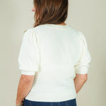 A woman viewed from behind wearing a cream-colored short-sleeve knit sweater with puffed sleeves and dark blue jeans, set against a plain light beige background.