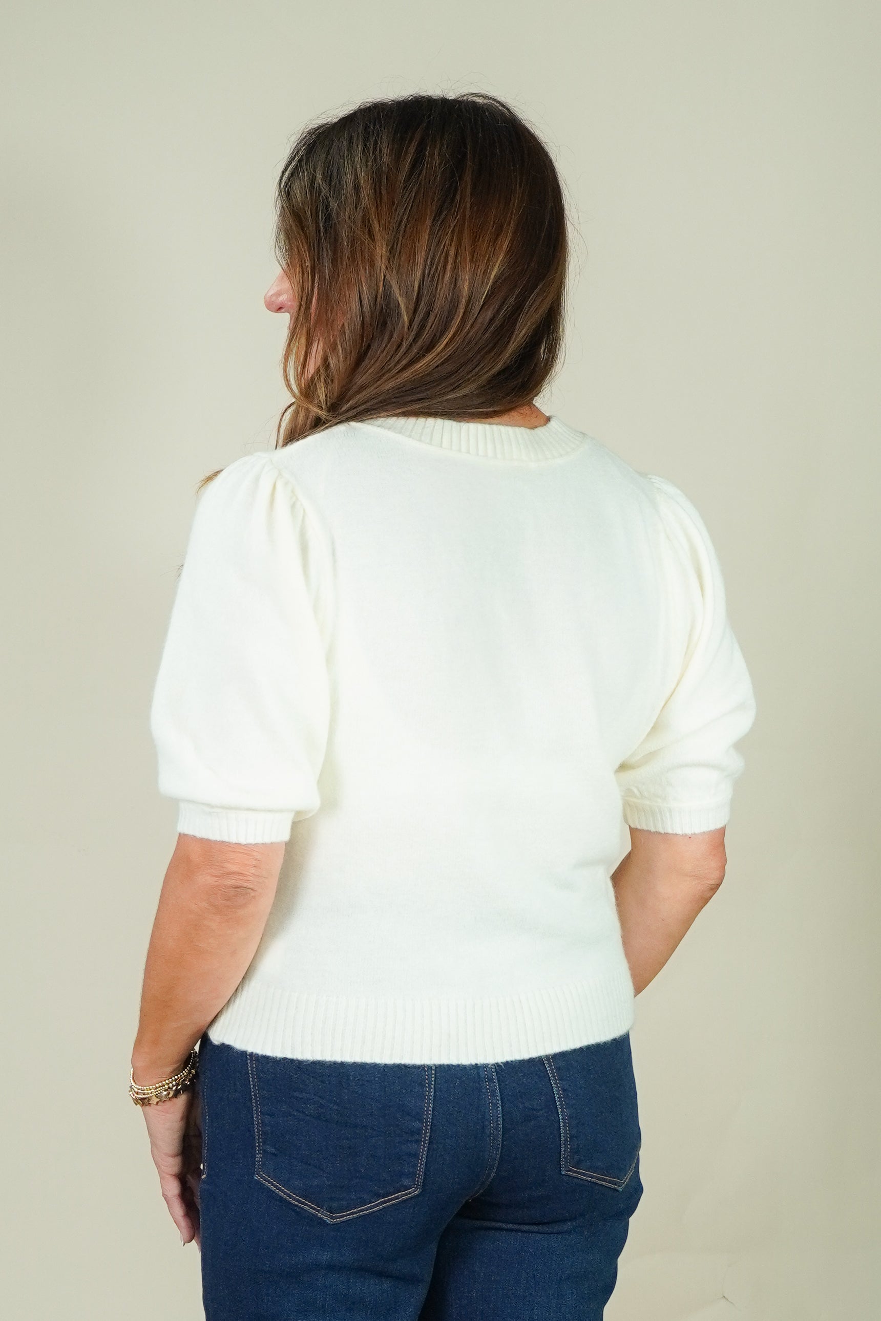 A woman viewed from behind wearing a cream-colored short-sleeve knit sweater with puffed sleeves and dark blue jeans, set against a plain light beige background.
