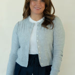 A woman wearing a light gray button-up cardigan over a white top and dark navy pants, standing against a plain off-white wall.