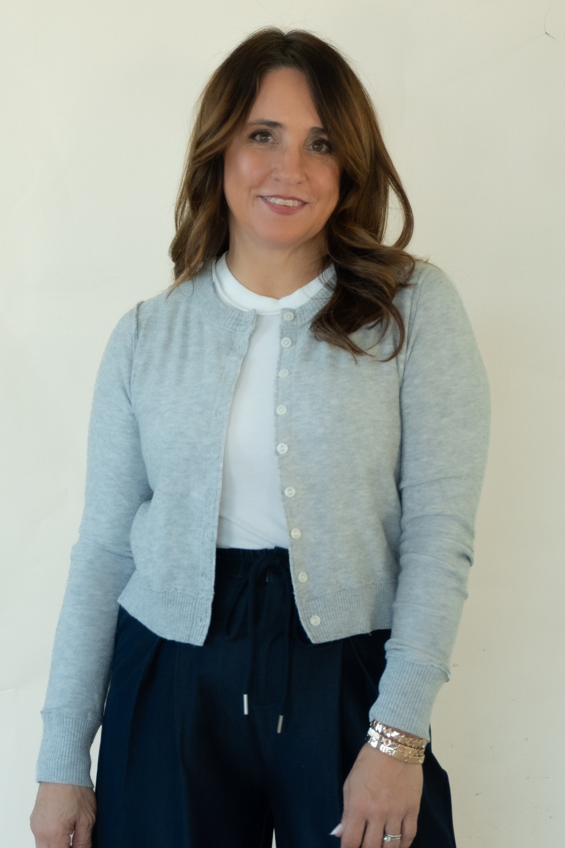 A woman wearing a light gray button-up cardigan over a white top and dark navy pants, standing against a plain off-white wall.