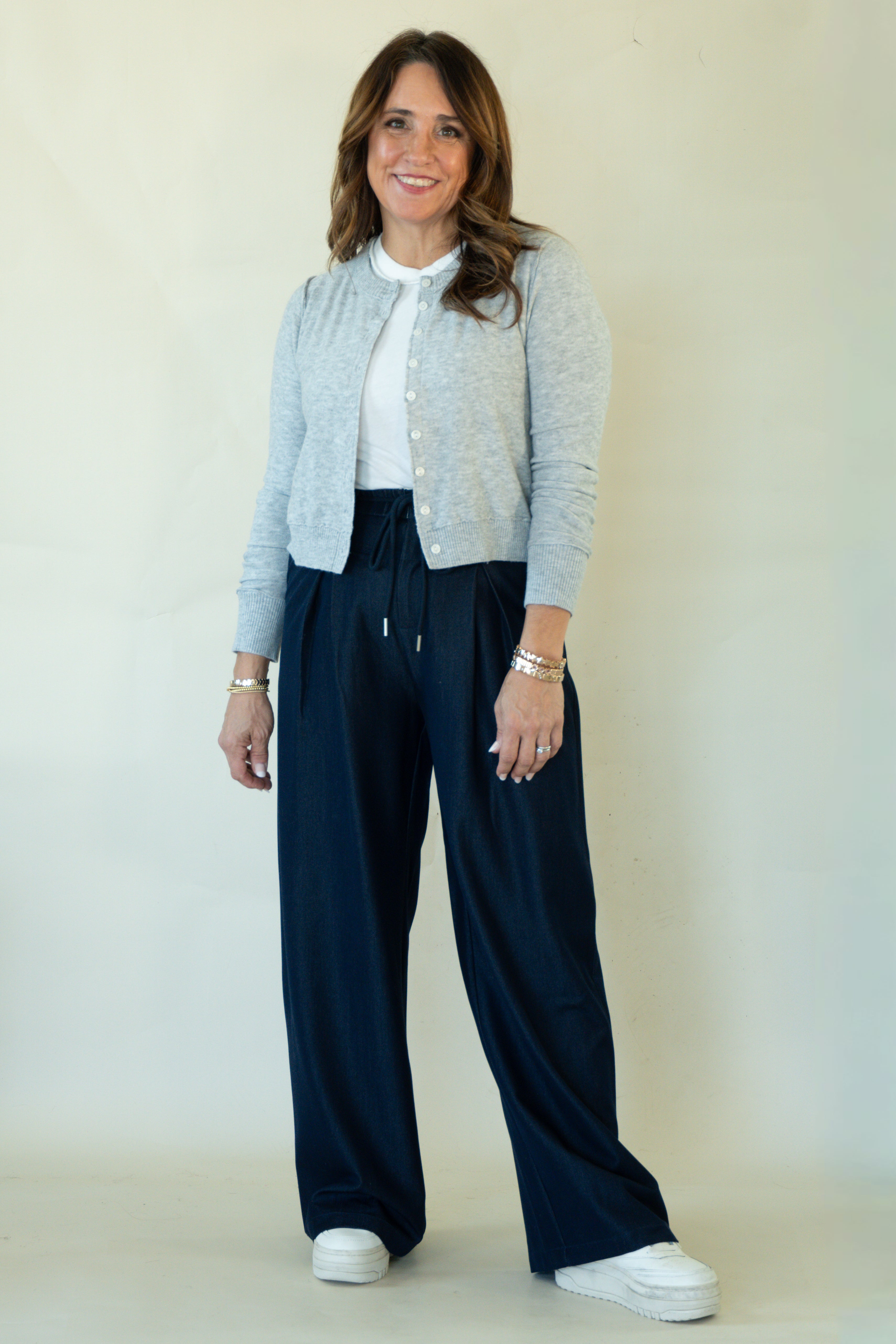 A woman modeling a light gray cardigan over a white top and navy wide-leg drawstring pants, standing against a plain off-white studio background.