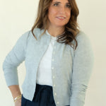 A woman wearing a light gray knit cardigan over a white top and dark navy pants, standing against a plain off-white background.