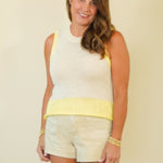 A woman modeling a sleeveless white knit top with a yellow waistband and matching white shorts, standing against a plain beige studio background.