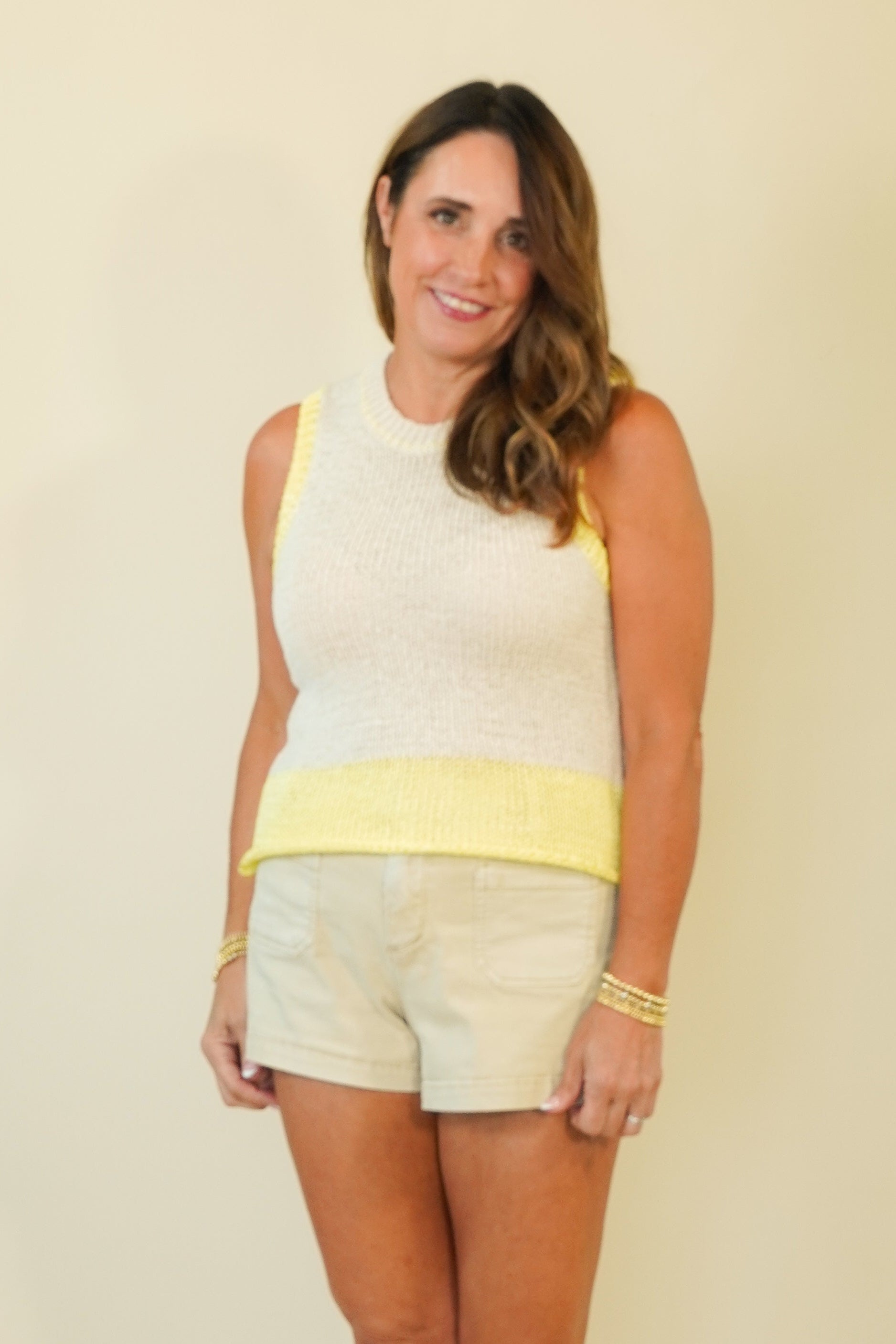 A woman modeling a sleeveless white knit top with a yellow waistband and matching white shorts, standing against a plain beige studio background.