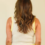 A woman viewed from behind wearing a sleeveless knit top with a light beige body and bright yellow trim along the armholes and waistband, against a plain cream background.