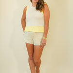 A woman modeling a sleeveless white knit top with a yellow waistband and white shorts, paired with white sandals, against a solid cream background.
