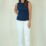 A woman modeling a sleeveless navy blue top and white cropped pants with beige wedge sandals, standing against a plain cream-colored studio background.
