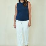 A woman modeling a sleeveless navy blue mock neck top and white cropped pants with beige sandals, standing against a plain cream-colored studio background.