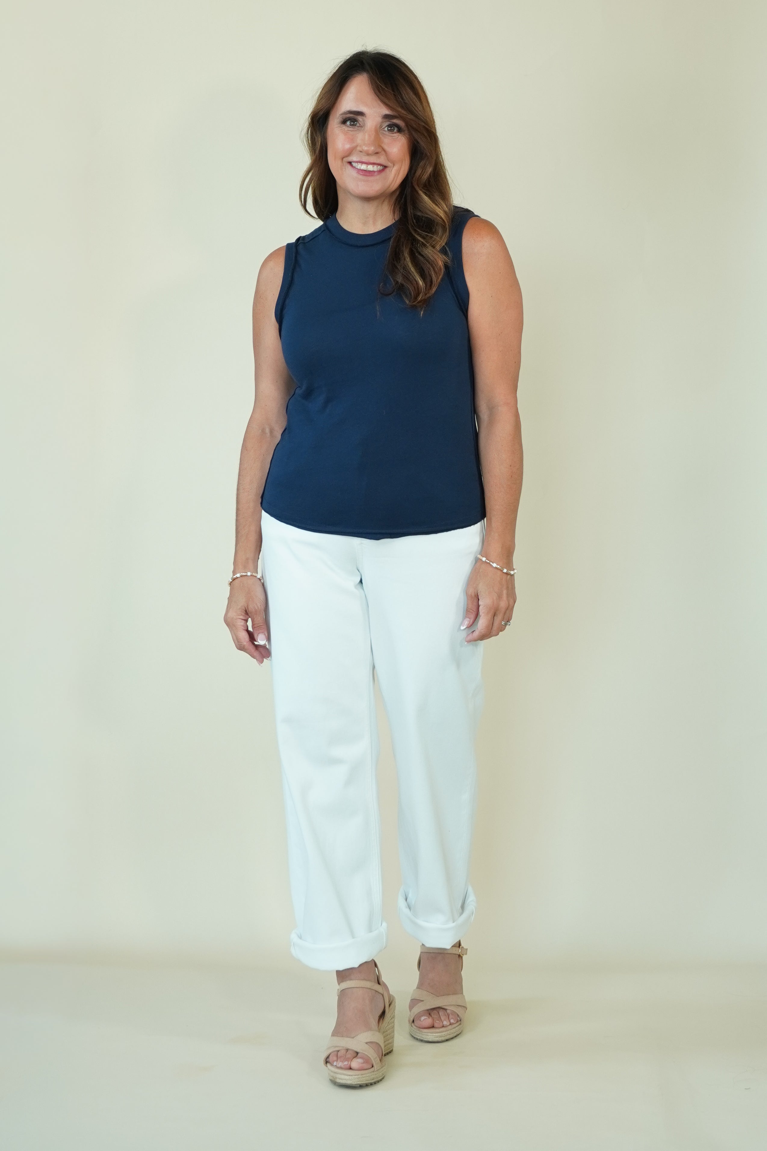 A woman modeling a sleeveless navy blue mock neck top and white cropped pants with beige sandals, standing against a plain cream-colored studio background.