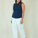 A woman modeling a sleeveless navy blue top and white cropped pants with beige wedge sandals, standing against a plain cream-colored studio background.
