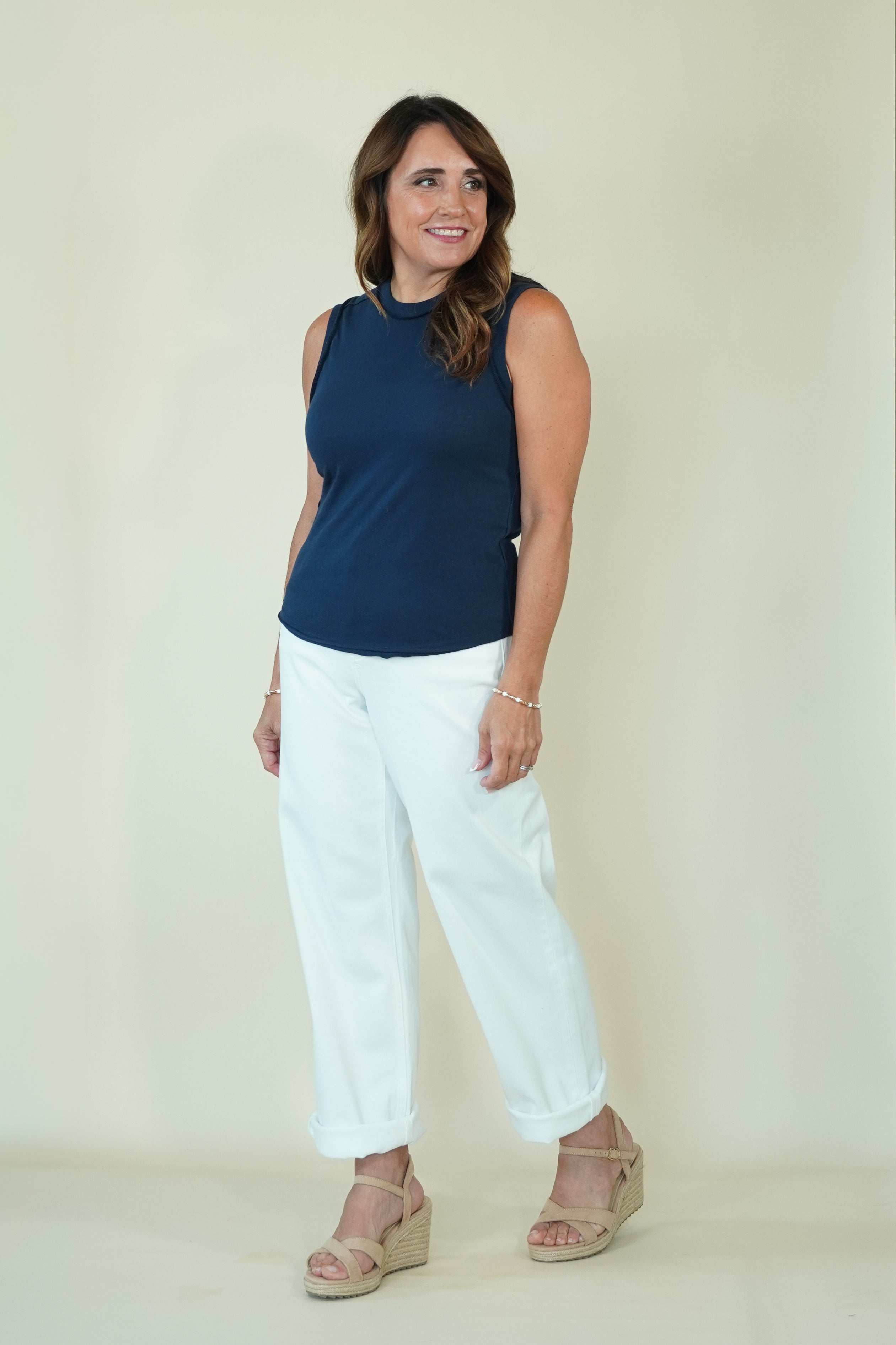 A woman modeling a sleeveless navy blue top and white cropped pants with beige wedge sandals, standing against a plain cream-colored studio background.