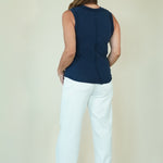 A woman shown from the back wearing a sleeveless navy blue top and white cropped pants with beige wedge sandals, standing against a plain light beige studio background.
