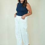 A woman modeling a sleeveless navy blue top and white cropped pants with beige wedge sandals, standing against a plain light beige studio background.
