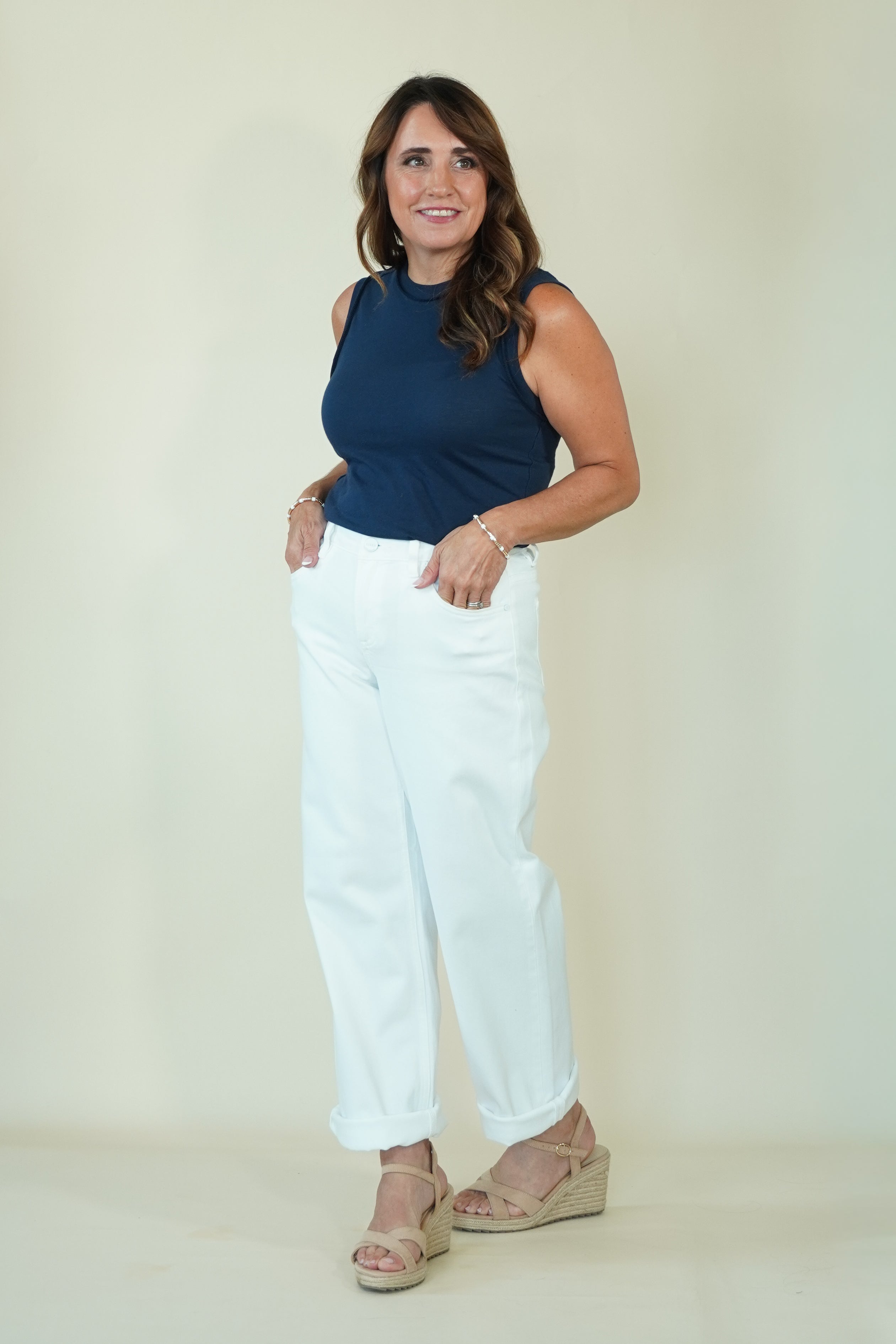 A woman modeling a sleeveless navy blue top and white cropped pants with beige wedge sandals, standing against a plain light beige studio background.