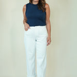 A woman modeling a sleeveless navy blue top and white cropped pants with beige wedge sandals, standing against a plain cream-colored studio background.