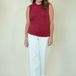 A woman modeling a sleeveless mock neck top in deep burgundy paired with white cropped pants and beige sandals, standing against a plain cream-colored studio background.