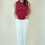 A woman modeling a sleeveless burgundy top and white pants against a plain cream background.