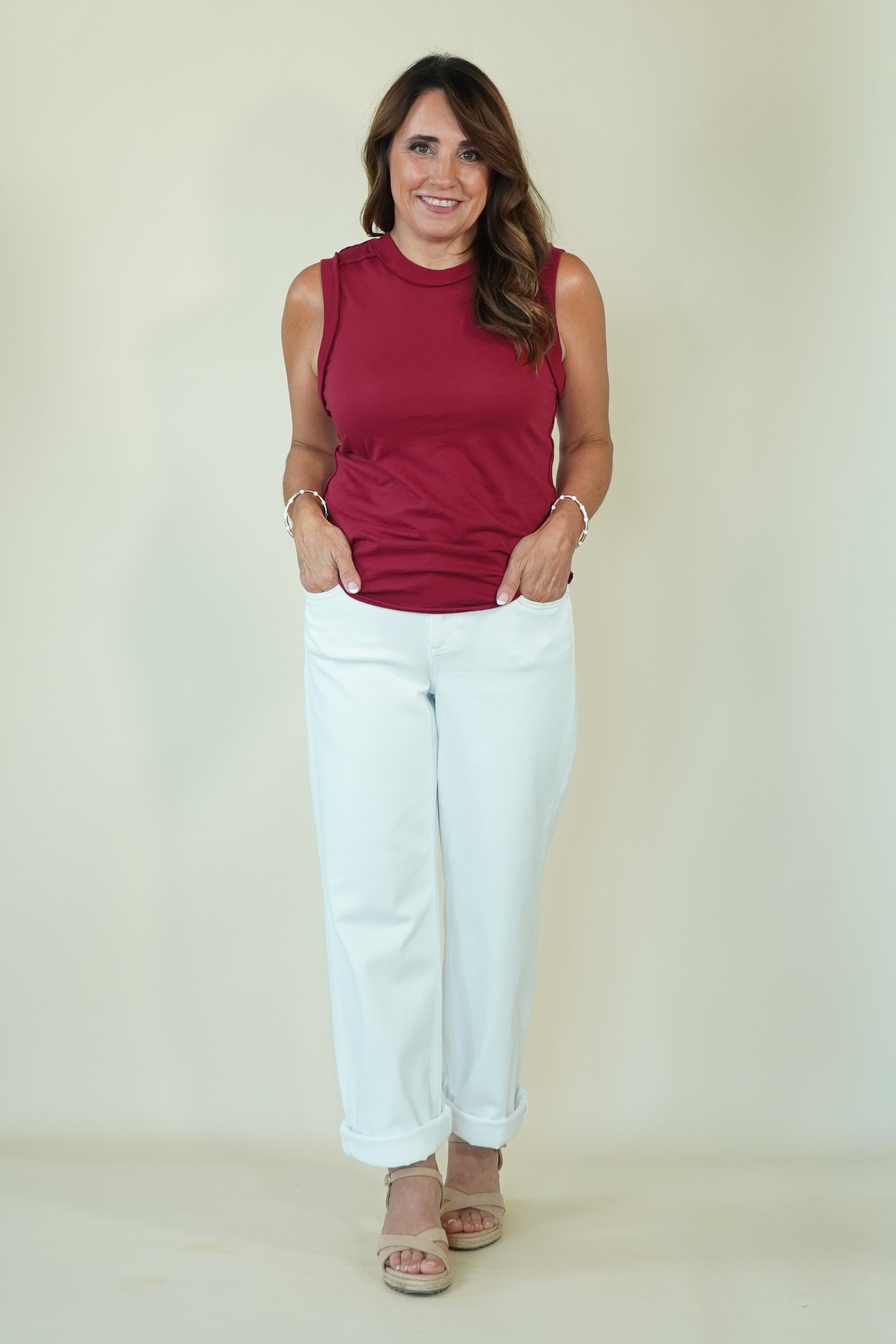 A woman modeling a sleeveless burgundy top and white pants against a plain cream background.