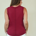 A woman viewed from the back wearing a sleeveless, scoop-neck knit top in deep burgundy with a vertical seam down the center back, paired with white pants, against a plain light beige background.