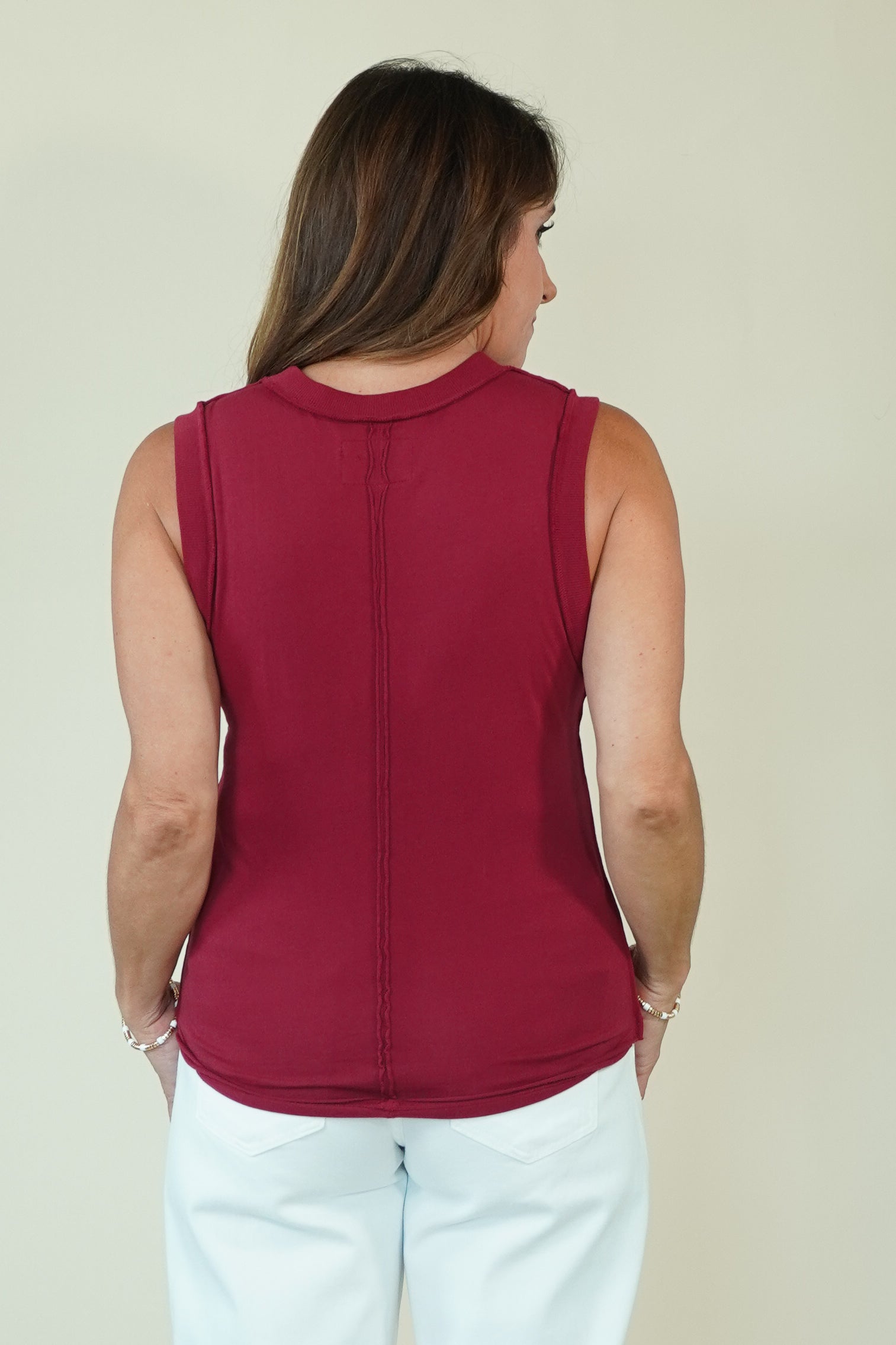 A woman viewed from the back wearing a sleeveless, scoop-neck knit top in deep burgundy with a vertical seam down the center back, paired with white pants, against a plain light beige background.