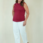 A woman modeling a sleeveless burgundy top and white cropped pants with beige wedge sandals, standing against a plain beige studio background.