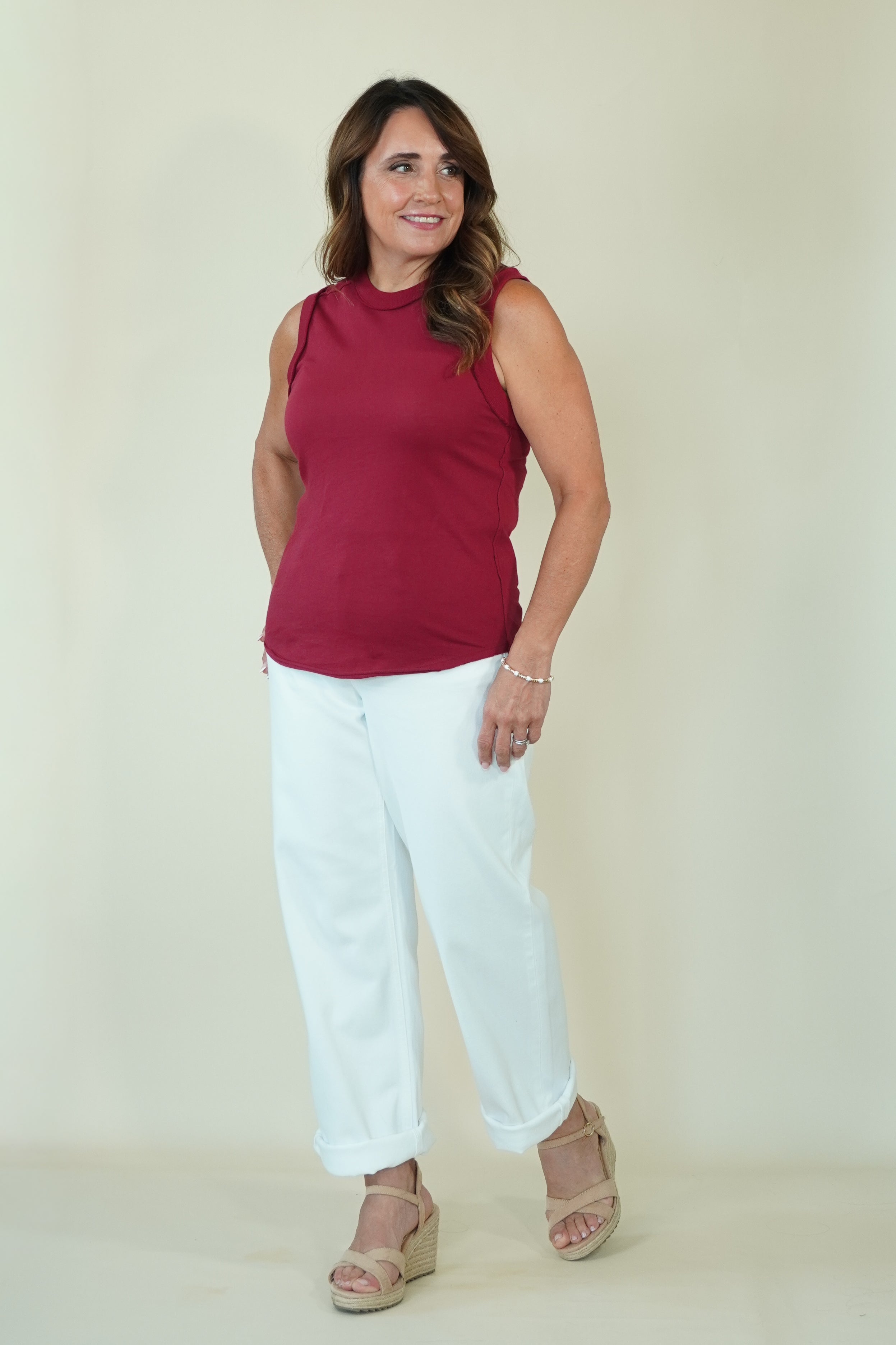 A woman modeling a sleeveless burgundy top and white cropped pants with beige wedge sandals, standing against a plain beige studio background.