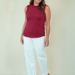 A woman modeling a sleeveless burgundy top and white cropped pants with beige wedge sandals, standing against a plain light beige studio background.
